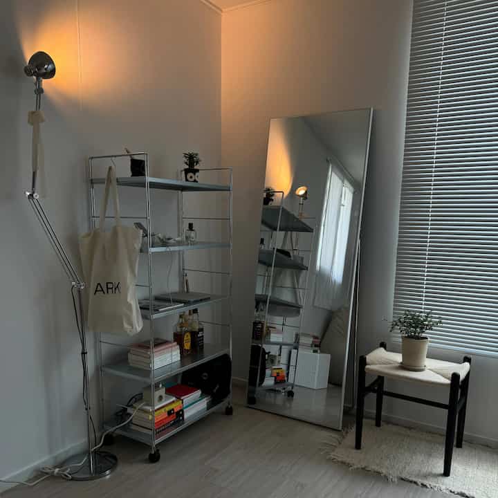白色のワンルーム空間、メタルの棚と大きな全身鏡、フロアランプとスツールが配置されたモダンで自然な雰囲気のインテリア