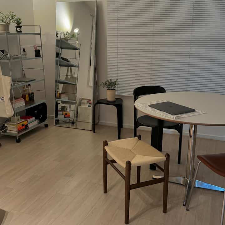 白い壁と木目調の床が調和する小さなダイニングルーム、丸テーブルとスツール、全身鏡が配置されたミニマルな空間