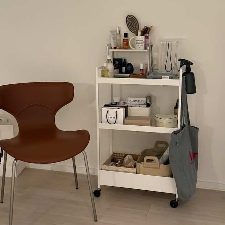 白い壁と床にブラウンレザーの椅子と化粧品を載せた白いトロリーが配置されたシンプルで実用的な一人暮らしの空間