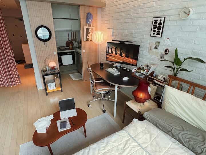 明るい木目調フローリングと白い壁の一人暮らし用ホームオフィス兼リビング空間、ミッドセンチュリーモダン家具とナチュラルモダン照明が配置された印象的なインテリア