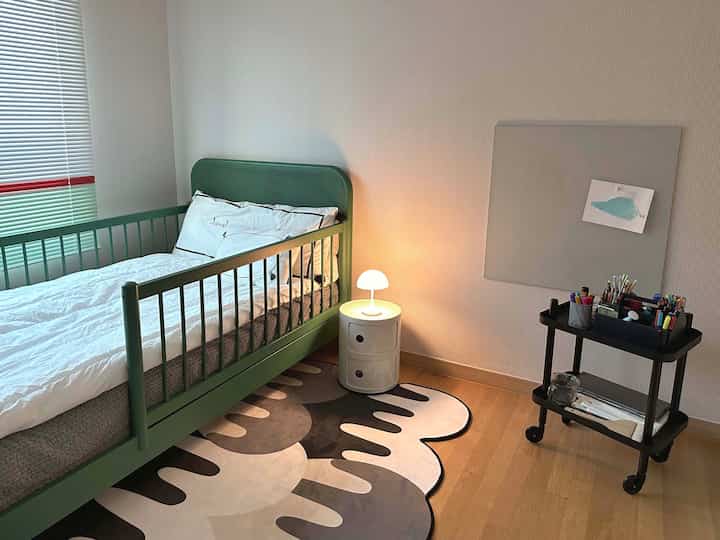 白い壁と緑の子供用ベッドが置かれたモダンな子供部屋、壁掛けボードと照明が特徴の空間