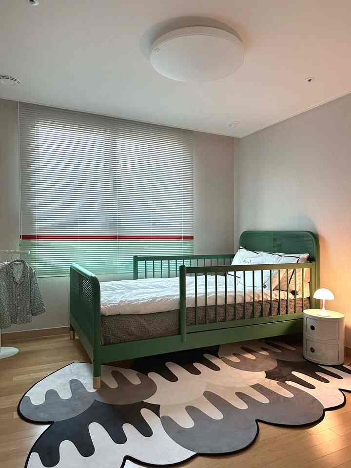 ナチュラルトーンの子供部屋にグリーンのベッドフレーム、独特なパターンのラグ、ブラインド、ナイトスタンドとテーブルランプが配置された落ち着いた雰囲気のインテリア
