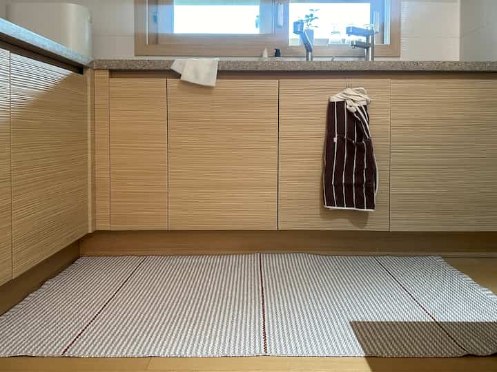 ベージュのウッド調が特徴のキッチン、床にPVC製のキッチンマットが敷かれ、シンクの水栓とキッチンタオルが配置された清潔感のある空間