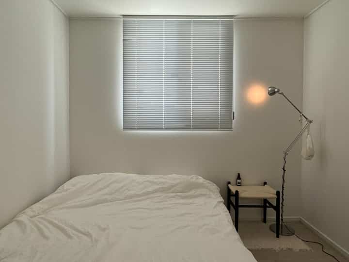 白とナチュラルカラーの寝室、ベッドとスツール、間接照明が配置された落ち着いた雰囲気のインテリア