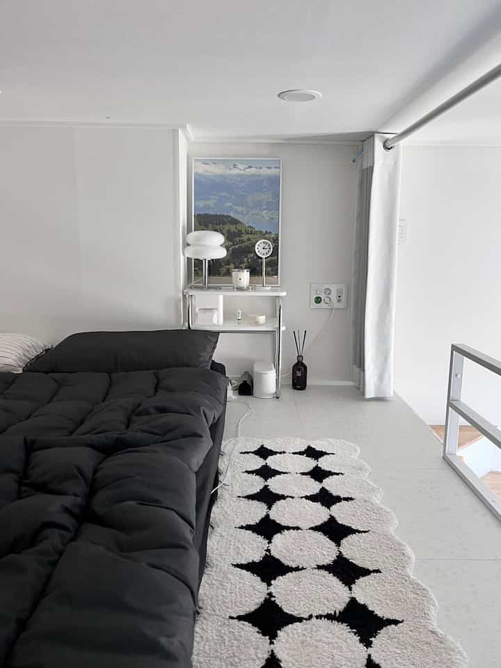 白と黒のモノトーンでまとめられた、低い天井の寝室。黒いベッド、白いナイトテーブル、風景のポスターが整然と配置された空間