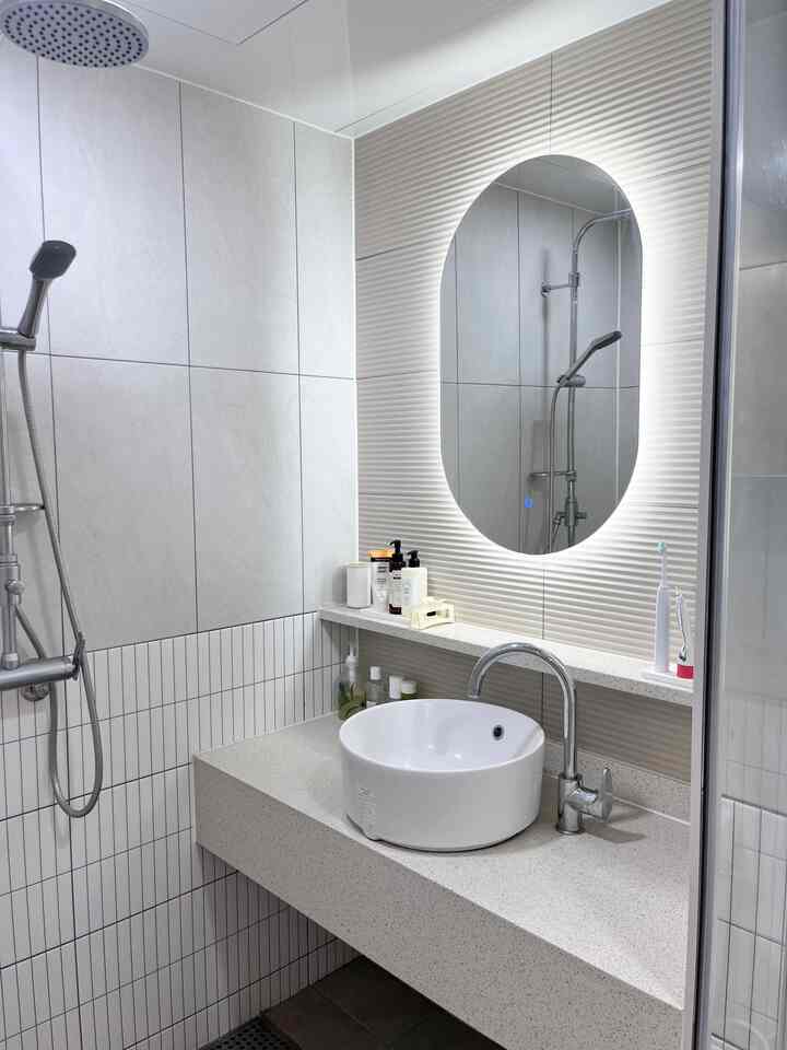 白を基調としたバスルーム、中央に丸型の洗面台とLED照明付きの楕円形の鏡が配置されたすっきりとした印象的な空間