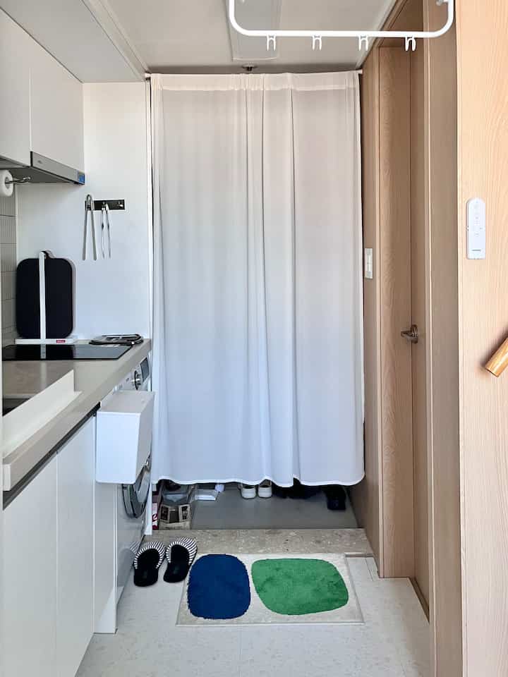 ホワイトとウッドトーンが調和した狭いキッチンと玄関、清潔感のあるカーテンとカラフルな玄関マットが配置された雰囲気のあるインテリア