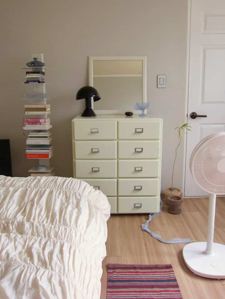 アイボリー調の寝室、ヴィンテージ風のチェストと白い寝具、テーブルランプが配置された雰囲気のある空間