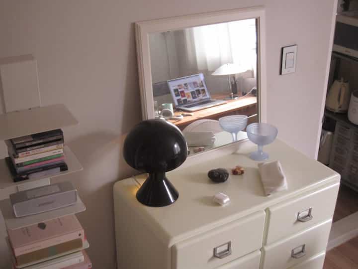 ホワイトとブラウンのトーンの狭い部屋、ドレッサーの上に鏡と黒い照明、鏡に映るデスクとテーブルランプがある化粧台