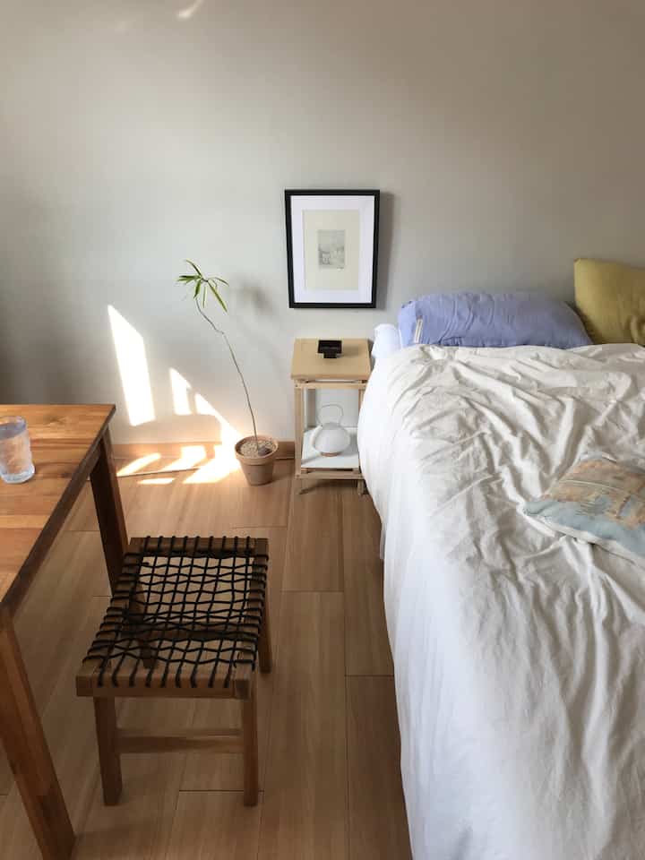 ナチュラルな木目調とホワイトが調和したワンルームの寝室、シンプルなベッドとナイトテーブル、植物が配置された落ち着いた雰囲気の空間