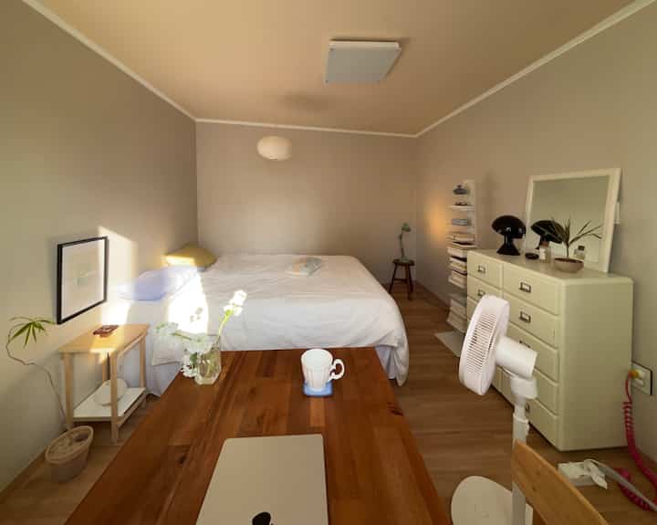 ベージュの壁と木目調の床が調和するモダンでナチュラルな寝室、白いベッドとチェストが配置された雰囲気のある空間