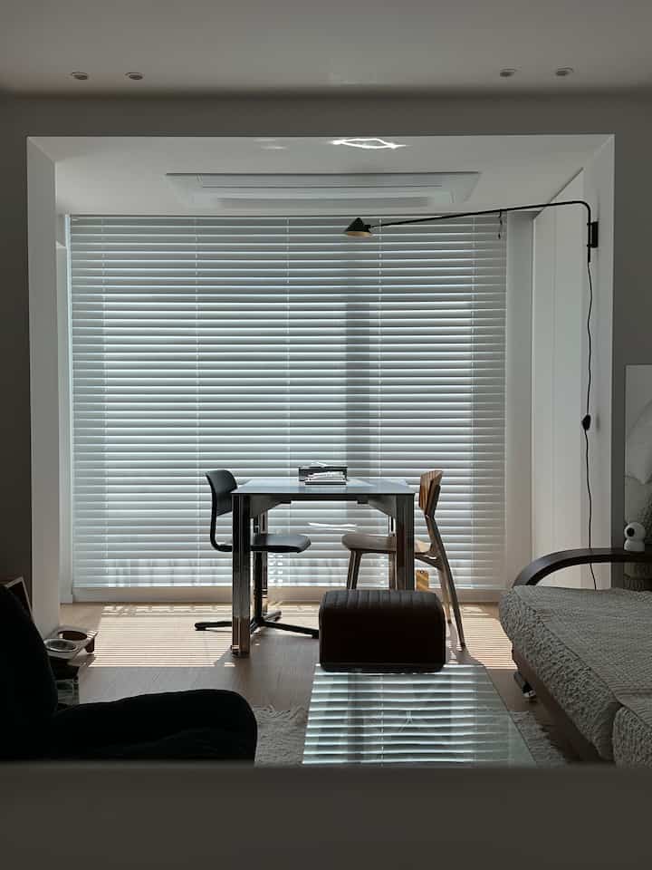 白とナチュラルカラーのリビング、ウッドブラインドと金属製テーブル、アームチェアとスツールが配置されたモダンな空間