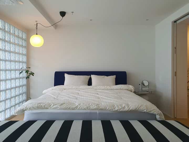 白と青のトーンの寝室、中央に大きなベッドと枕、左側にガラスブロックの壁とペンダント照明が配置されたシンプルな空間