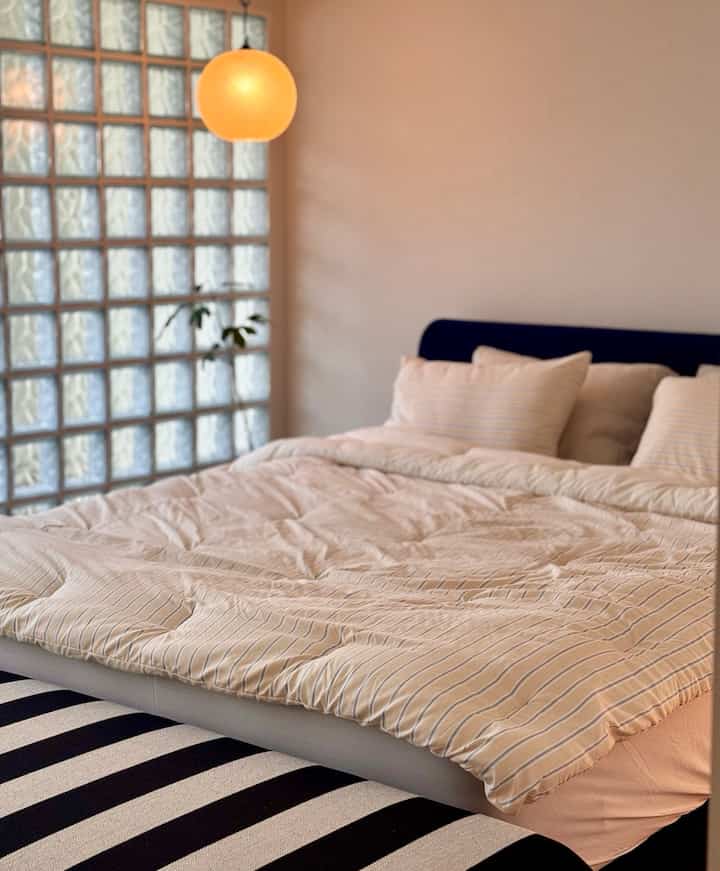 温かみのあるアイボリー色調の寝室、ネイビーブルーのベッドヘッドとストライプ柄のベンチが配置されたナチュラルモダンな空間