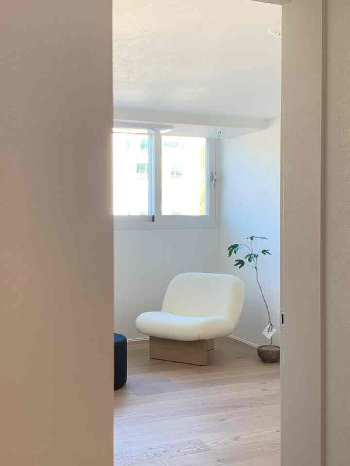 明るいホワイトとナチュラルな木目調の狭い部屋、窓辺に配置された白いラウンジチェアと植物がある思索の空間