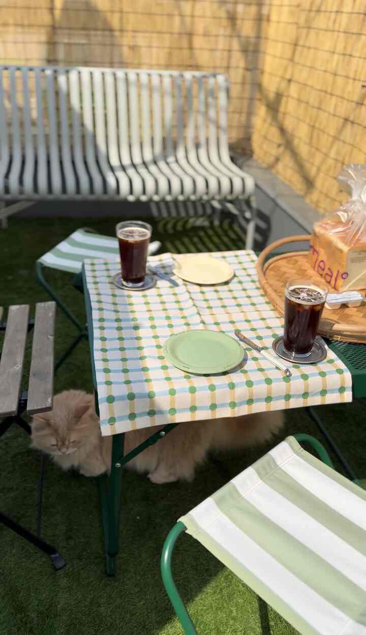 緑の人工芝の床が敷かれたテラスに、緑と白の屋外テーブルとチェアが配置され、猫とアイスコーヒーがある居心地の良いおうちカフェのインテリア