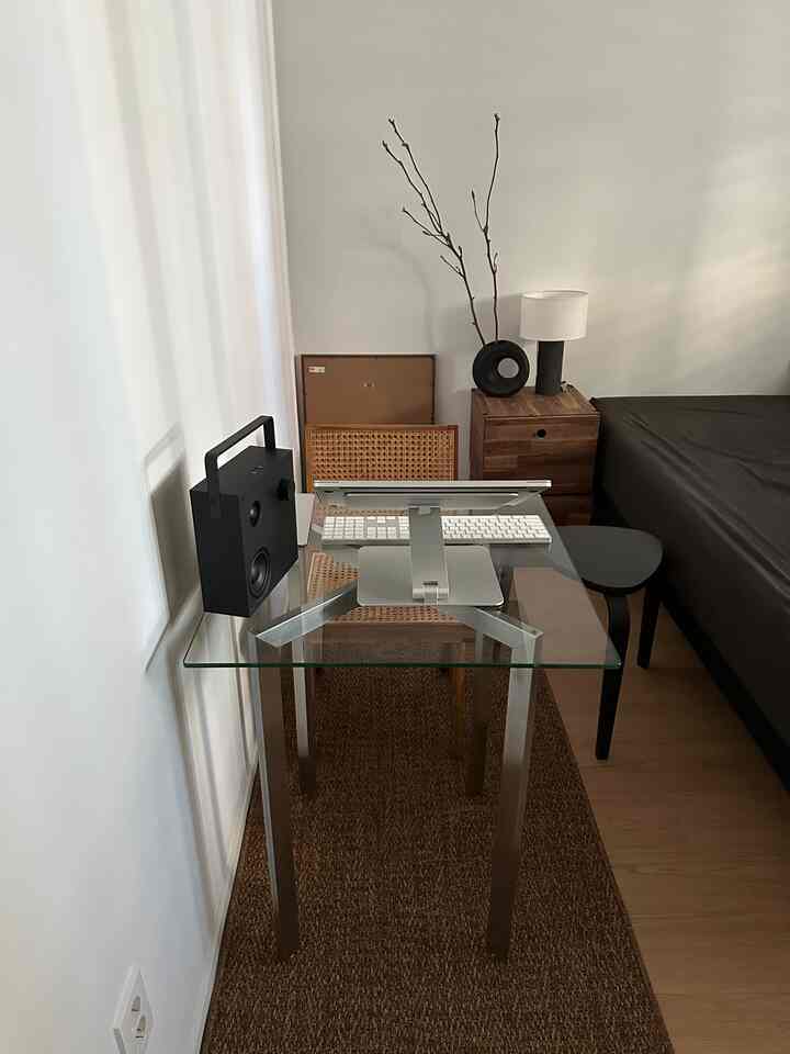 ブラックとナチュラルブラウンのトーンの寝室兼ホームオフィス、ガラステーブルとナイトテーブル、ラグが配置されたモダンで居心地の良い空間