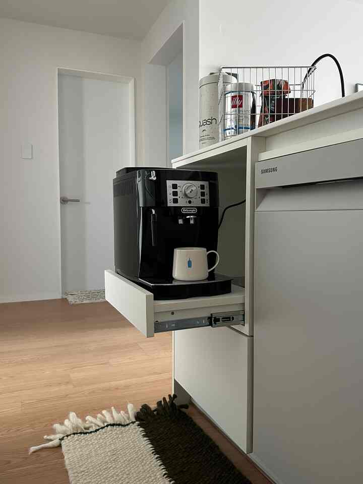 ホワイトとブラウンの色調のキッチンスペース、自動コーヒーマシンとマグカップ、ラグが配されたシンプルでモダンなホームカフェのインテリア