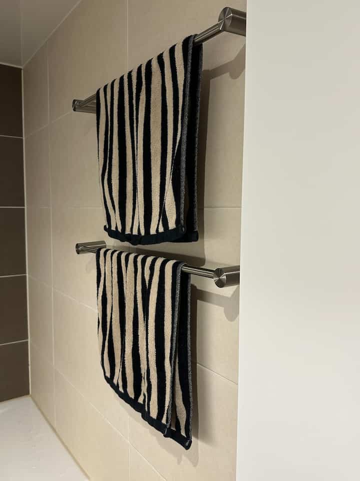 ベージュのタイル壁に金属製のタオル掛けがあり、黒とベージュのストライプタオルが掛けられたバスルームのインテリア