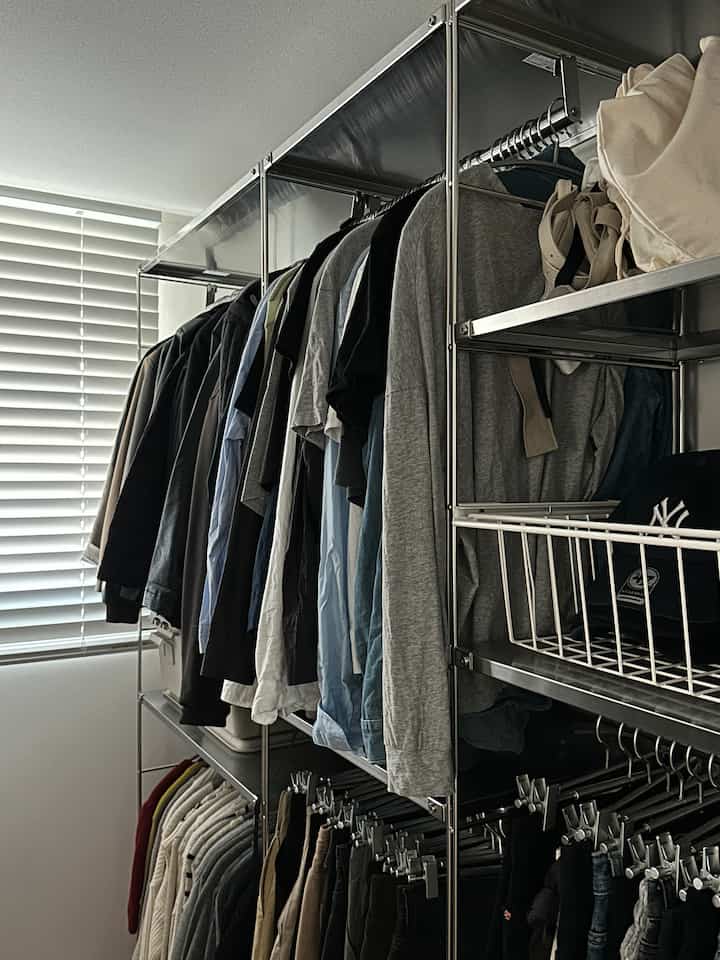 グレーの衣服が掛けられた金属製クローゼットが配置されたワンルームの内部、白いブラインドが窓を覆っている