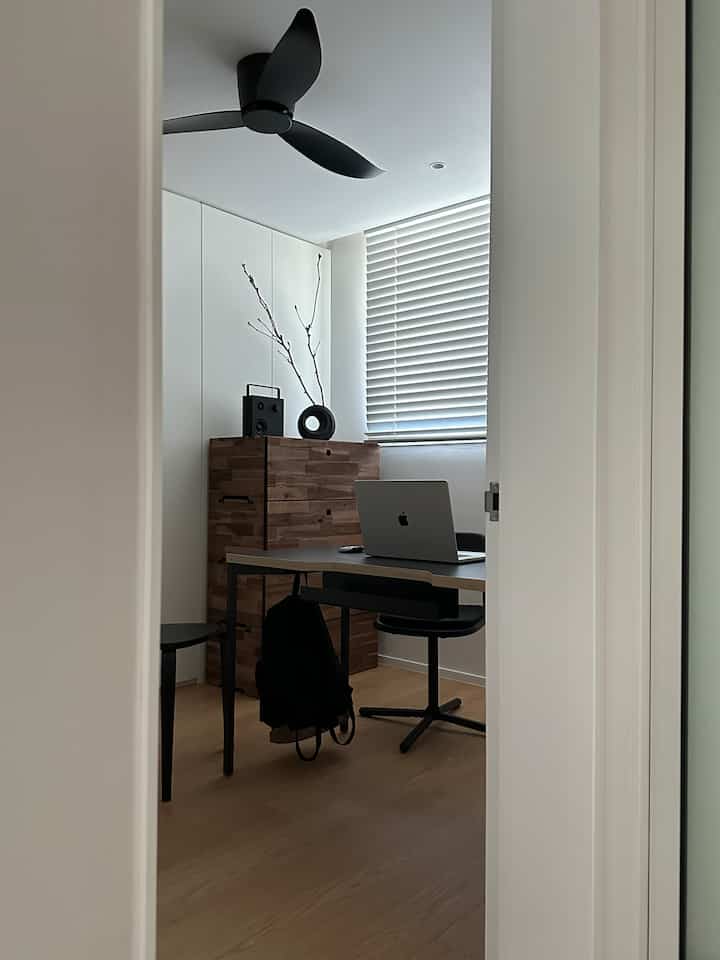 白と木目調の小さなホームオフィス、デスクとオフィスチェア、クローゼットとシーリングファンが配置された清潔感のある空間