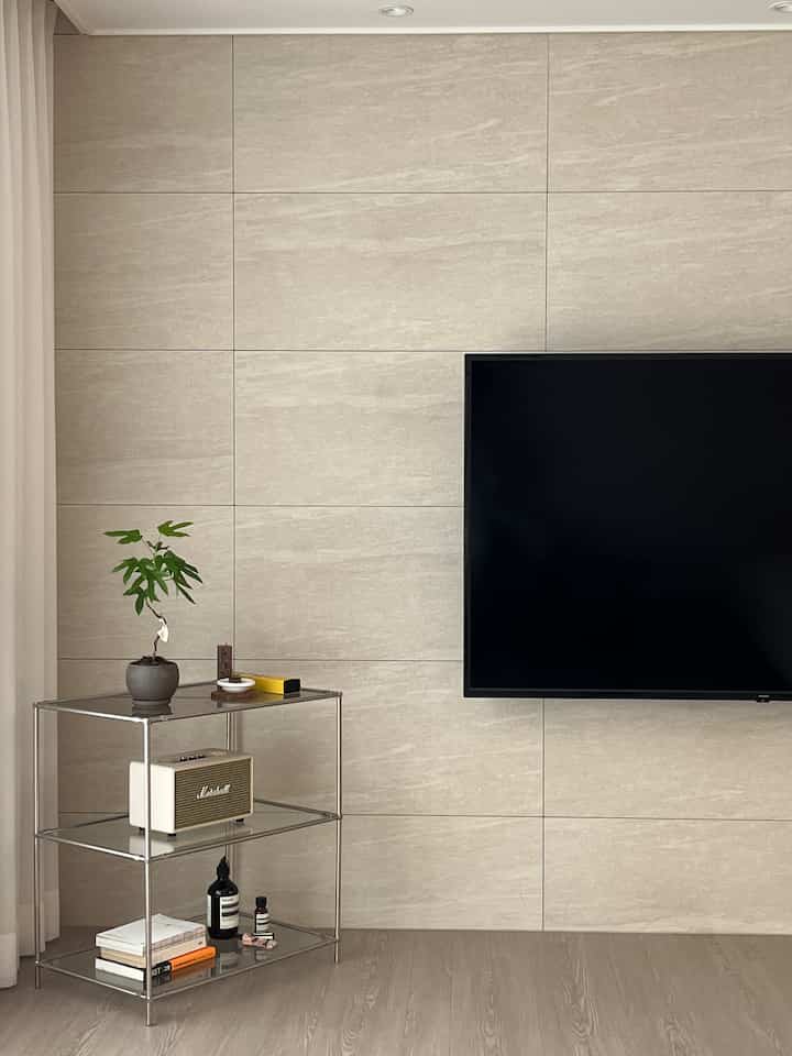 ベージュの壁とナチュラルトーンの床のリビング、メタルシェルフとテレビが配置されたモダンでミニマルな空間
