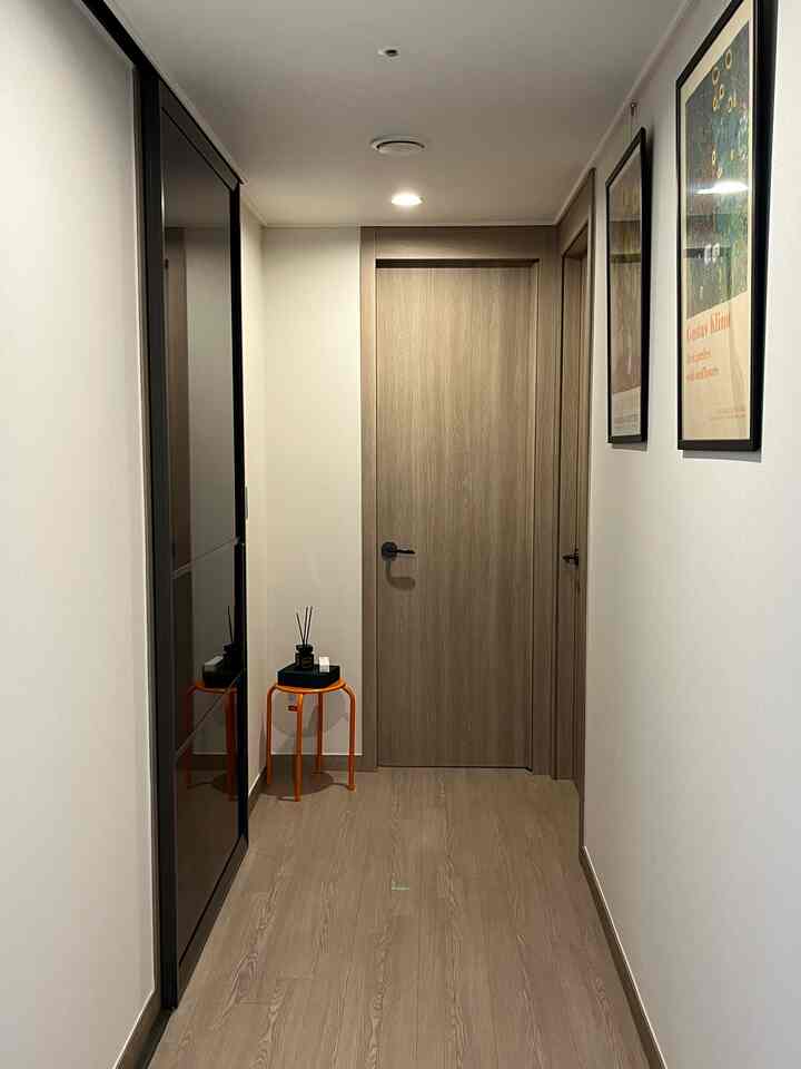 白い壁と木目調の床とドアがある廊下に、オレンジ色のスツールとディフューザー、右側の壁に掛けられた2つの絵画が配置されたシンプルなインテリア