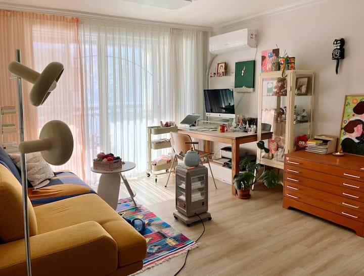 白いカーテンとナチュラル調の家具が調和するリビング兼ホームオフィス、黄色のソファとラグが暖かな雰囲気を演出