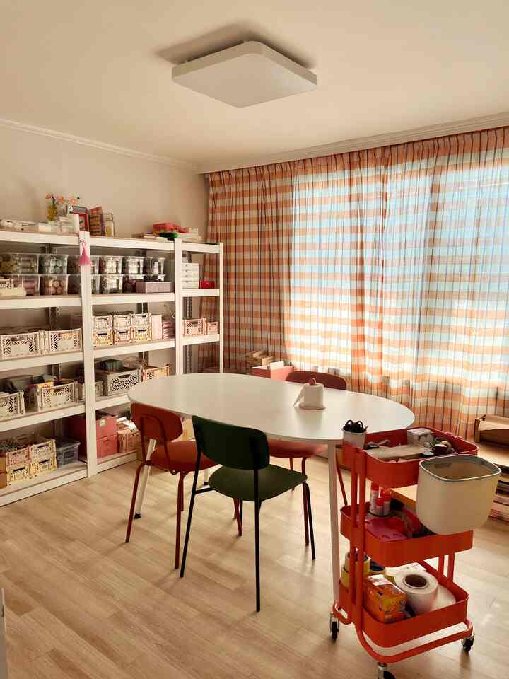 暖かいオレンジのチェック柄カーテンと木目調の床がある小さなダイニングルーム、白い楕円形のダイニングテーブルとカラフルな椅子が配置された整理された空間