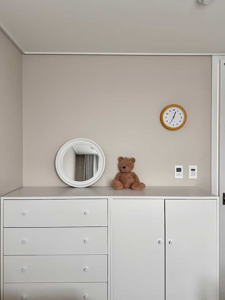 ベージュの壁とホワイトのチェストがある赤ちゃん部屋、ブラウンのテディベアと丸い時計がアクセントの清潔感のあるインテリア