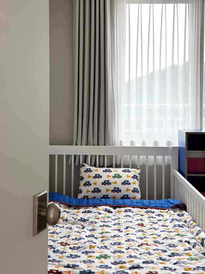 白と青のトーンの子供部屋、白いベッドフレームのベビーベッドとかわいい車柄の寝具が配置された印象的な空間
