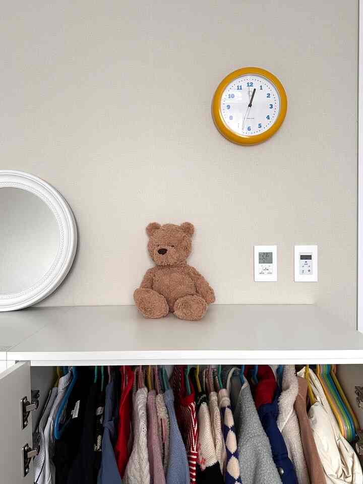 ナチュラルなベージュと白のベビールームのクローゼットが中央に配置され、壁に黄色い時計と茶色のテディベアが飾られた印象的なインテリア