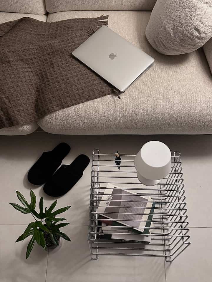 ナチュラルカラーのリビング、ベージュのソファにブラウンのブランケット、ワイヤーキューブテーブル上にノートパソコンとテーブルランプが配置された雰囲気のある空間