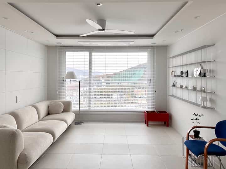 白を基調としたリビング、シンプルなソファとブラインドの窓、シーリングファンと木目調の椅子が配置された清潔感のあるインテリア