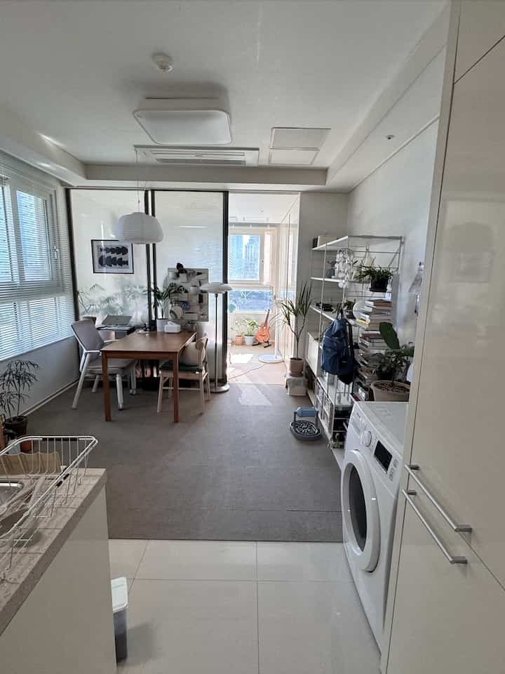 白と茶色のトーンのリビング兼ホームオフィス空間、ダイニングテーブルとデスク、多様な植物が配置された雰囲気のあるインテリア