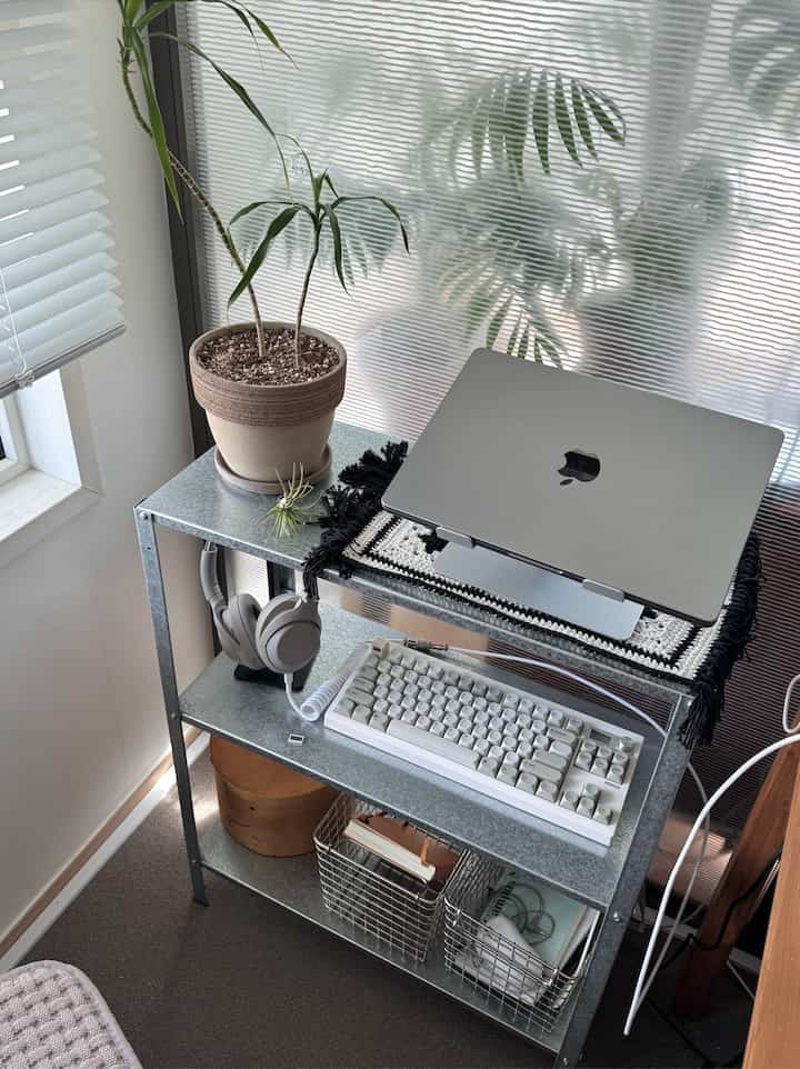 シルバーの金属棚と白いキーボードがあるコンパクトな在宅勤務スペース、ブラインドと植物が配置されて整った雰囲気のインテリア
