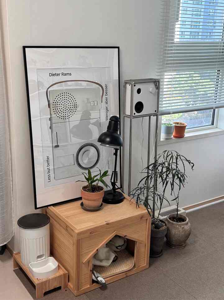 白いブラインドの窓の横に木製のペットハウスと複数の鉢植えが配置されたナチュラルトーンの空間