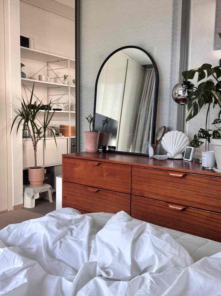 白い寝具とナチュラルウッドのチェストがある寝室、アーチ型の鏡と植物が配置された雰囲気のあるインテリア