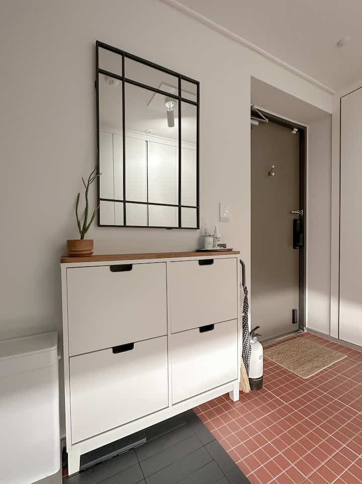 アイボリー色の4つのコンパートメントの靴棚と黒い格子の壁鏡、赤いタイルの床がある整然とした玄関スペース