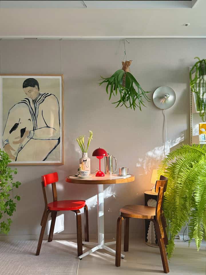 緑の植物が豊富な寝室のカフェ風ダイニングスペース、赤い椅子とテーブルランプがアクセントの温かみのあるインテリア