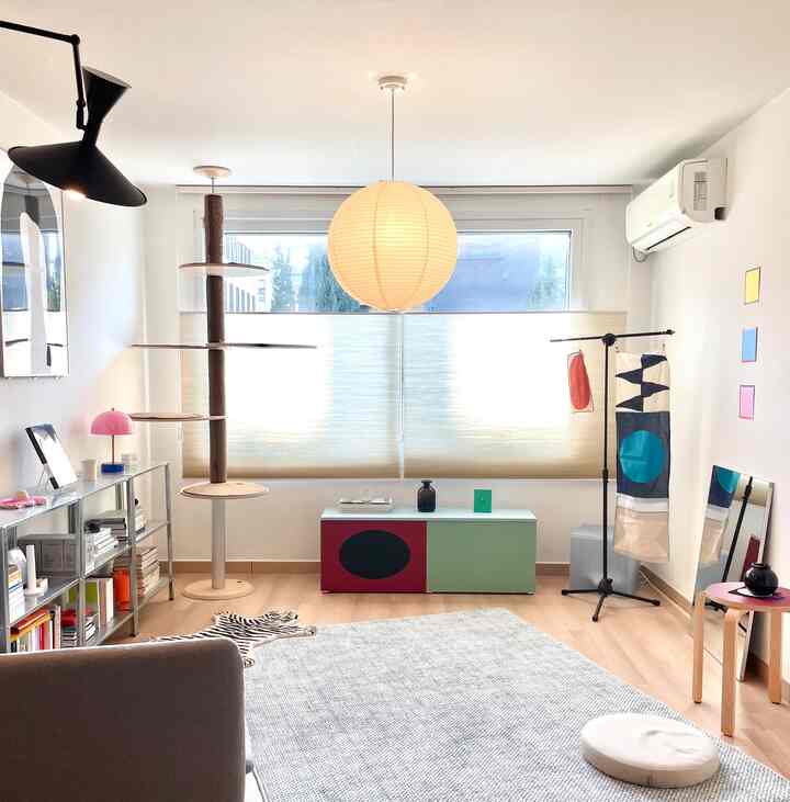 白い壁と木目調の床のナチュラルなリビング、ハニカムブラインドが窓を覆い、キャットタワーとラグが配置された雰囲気のあるインテリア