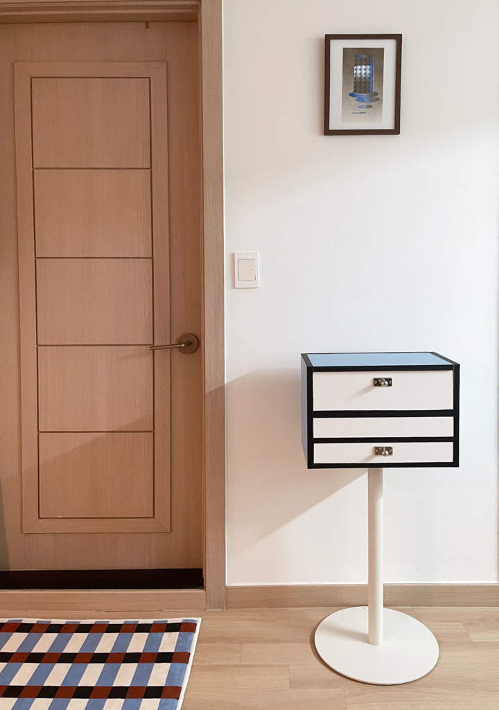 白い壁と茶色の木製ドアが調和する空間に、白と黒のナイトテーブルが置かれたモダンなリビングインテリア