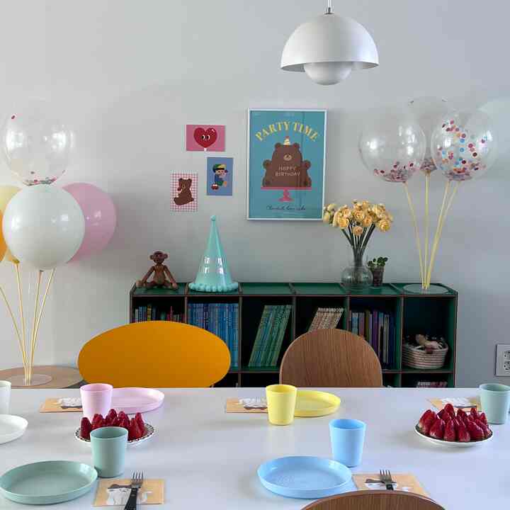 白い背景の子供部屋、緑の本棚にパーティーポスターと風船、パステルカラーの食器と椅子が配置された雰囲気のあるホームパーティースペース