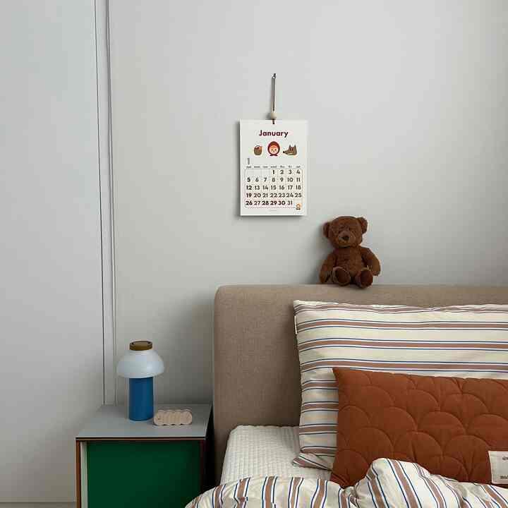 ベージュとブラウンの色調の子供部屋、ベッドにぬいぐるみ、壁にかけられた赤ずきんをテーマにしたカレンダーがある印象的な空間