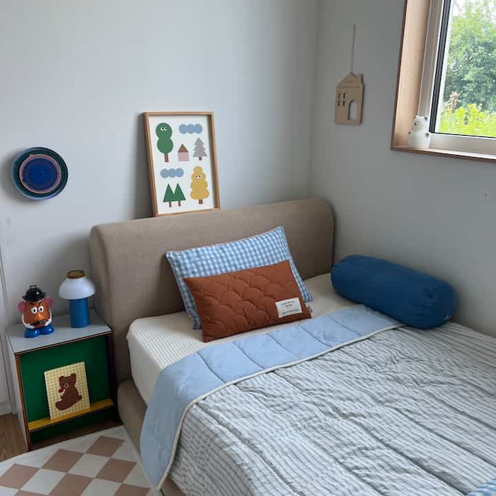 白い壁とナチュラルな木目調の子供部屋、ベージュのベッドとパステルブルーの寝具、かわいらしいポスターとクッションが配置された落ち着いた空間