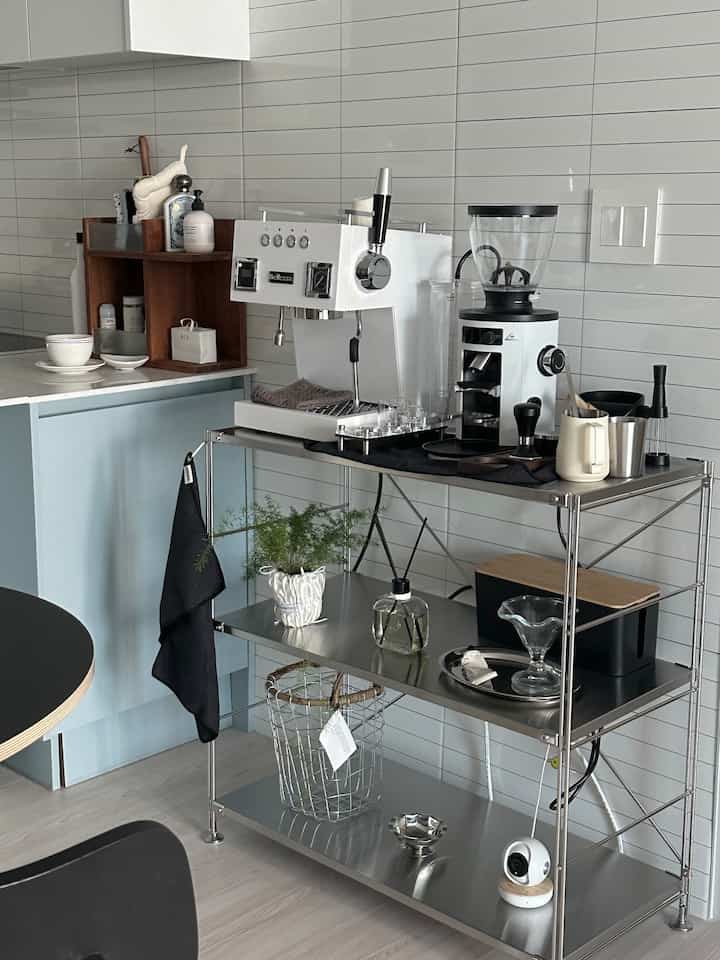 ホワイトとシルバーのトーンのキッチンのおうちカフェ、ステンレス棚にコーヒーマシンやコーヒー用品が整ったモダンなインテリア