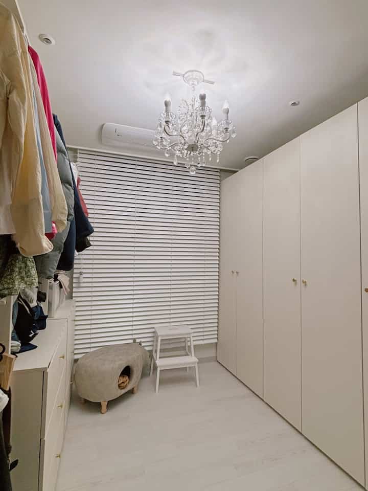 明るいホワイトトーンのウォークインクローゼット、ホワイトのビルトイン収納と引き出し、横型ウッドブラインド、猫のためのグレーのペットハウスが配置された清潔感のあるシンプルな空間