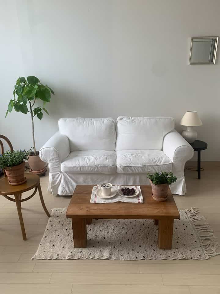 白とナチュラルウッドトーンが調和するリビング、二人掛けの白いソファと植物とコーヒーのある木製コーヒーテーブルが配置された落ち着いた空間