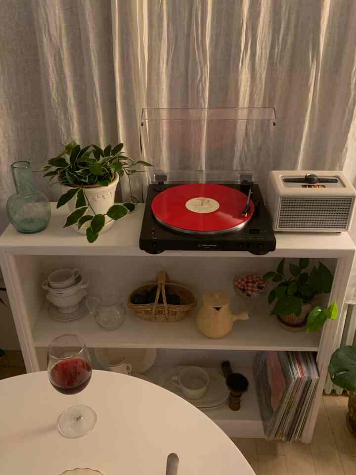 温かみのあるベージュのリネンカーテンを背景に、白いキャビネットと赤いLPターンテーブル、さまざまなオブジェと植物が配置された雰囲気のあるインテリア