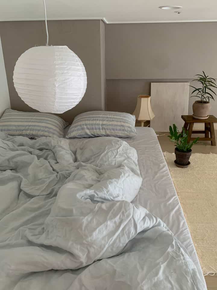 ベージュ色の壁と木製スツール、植物が配された落ち着いた寝室に青いストライプの寝具が置かれた空間